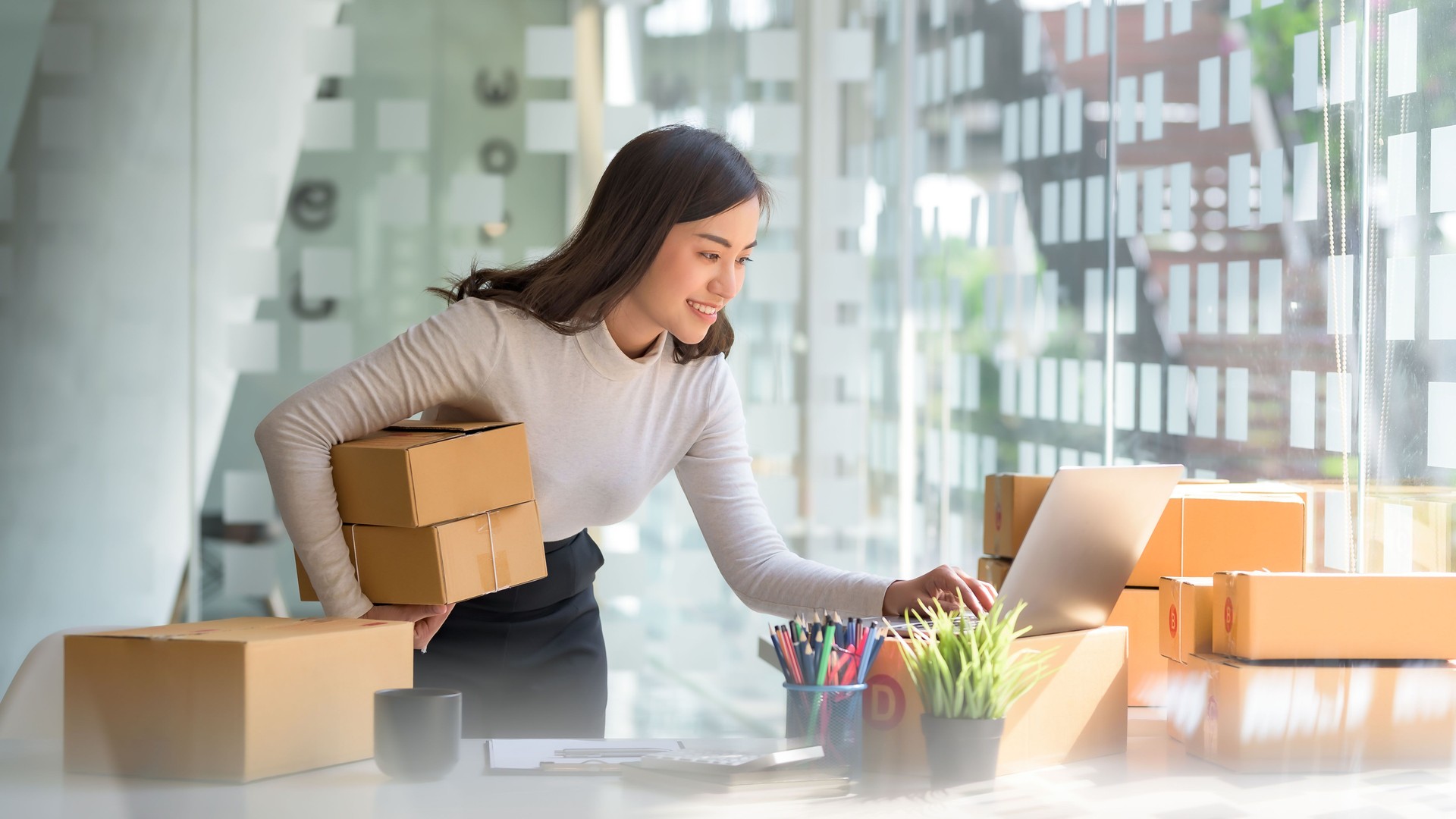 Smiling young Asian business owner woman prepare parcel box and standing check online orders of product for deliver to customer on laptop computer. Shopping Online concept.