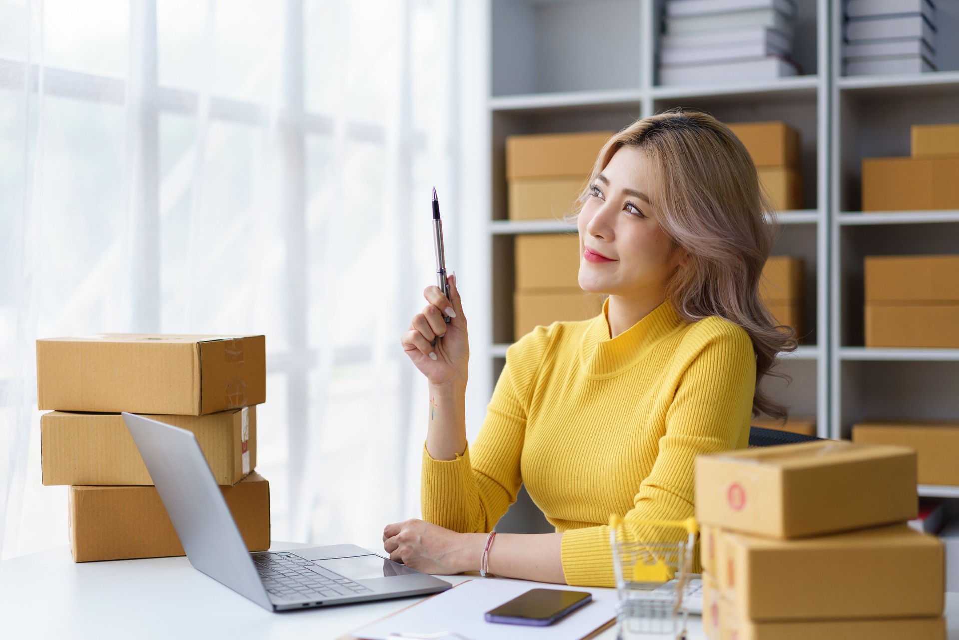 Thoughtful young businesswoman planning strategy surrounded by shipping boxes in cozy home-based online store